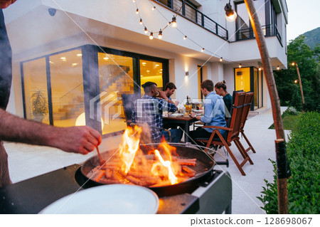 A group of friends and family barbecue together in the evening on the terrace in front of a large modern house. A group of friends and family barbecue together in the evening on the terrace in front of a large modern house. 128698067