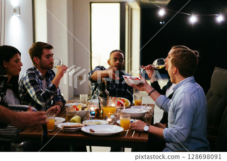 A group of young diverse people having dinner on the terrace of a modern house in the evening. Fun for friends and family. Celebration of holidays, weddings with barbecue 128698091
