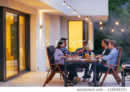 A group of young diverse people having dinner on the terrace of a modern house in the evening. Fun for friends and family. Celebration of holidays, weddings with barbecue 128698101