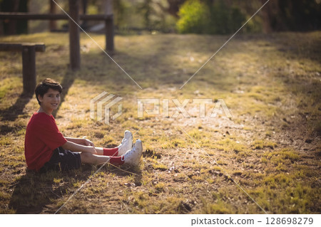 Smiling boy sitting on grass in park, enjoying sunny day outdoors, copy space 128698279