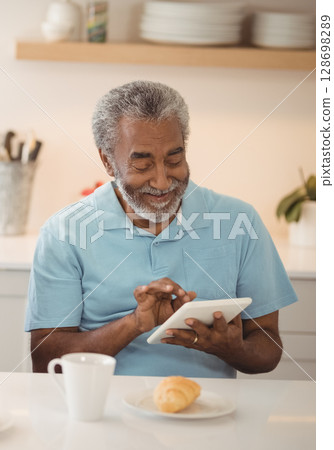 Elderly man enjoying breakfast while using tablet at home kitchen table 128698289