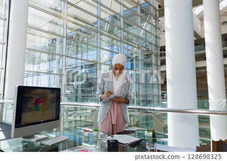 Businesswoman taking notes in modern office with computer and glass walls Businesswoman taking notes in modern office with computer and glass walls 128698325