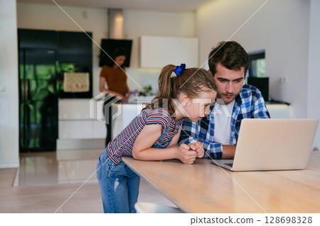Work from home. Daughter interrupting her father while he is having a business online conversation on his laptop while sitting in the modern living room. Work from home. Daughter interrupting her father while he is having a business online conversation on his laptop while sitting in the modern living room. 128698328