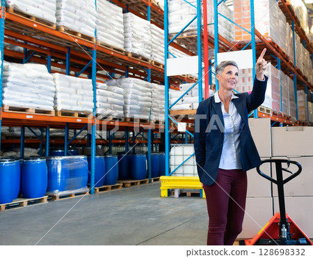 In warehouse, businesswoman pointing while managing inventory with pallet jack, copy space In warehouse, businesswoman pointing while managing inventory with pallet jack, copy space 128698332