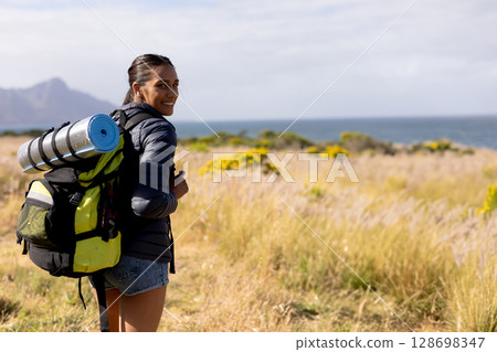 Smiling woman hiking with backpack and yoga mat in scenic nature, copy space 128698347