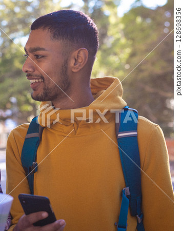 Smiling young man in yellow hoodie holding smartphone outdoors with backpack 128698356