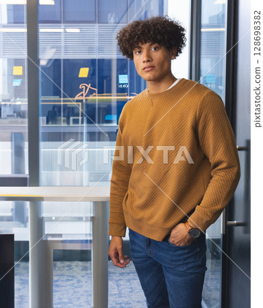 Confident young man standing by office window, wearing casual sweater and jeans Confident young man standing by office window, wearing casual sweater and jeans 128698382