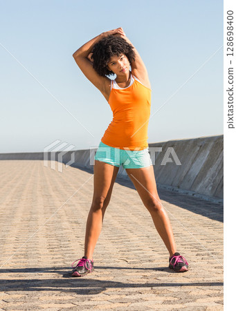Woman stretching outdoors on sunny promenade, wearing bright athletic outfit 128698400