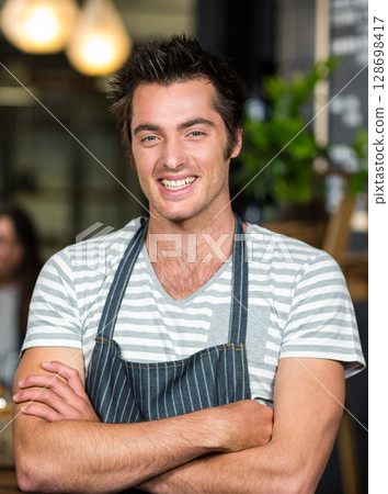 Barista smiling confidently in coffee shop, wearing striped apron, arms crossed 128698417