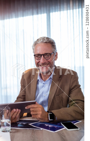 Smiling businessman using tablet at office desk with smartphone and documents Smiling businessman using tablet at office desk with smartphone and documents 128698431