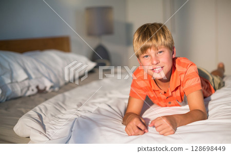 Smiling boy in orange shirt relaxing on bed at home, enjoying leisure time, copy space 128698449