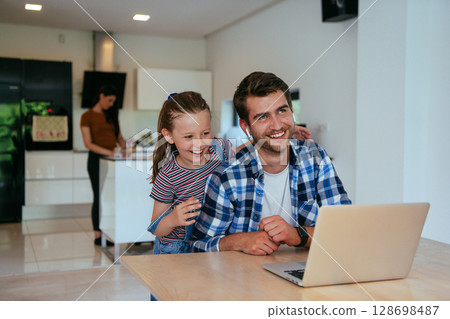 Work from home. Daughter interrupting her father while he is having a business online conversation on his laptop while sitting in the modern living room. Work from home. Daughter interrupting her father while he is having a business online conversation on his laptop while sitting in the modern living room. 128698487