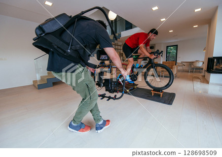 A cameraman filming an athlete riding a triathlon bike on a simulation machine in a modern living room. Training in pandemic conditions A cameraman filming an athlete riding a triathlon bike on a simulation machine in a modern living room. Training in pandemic conditions 128698509