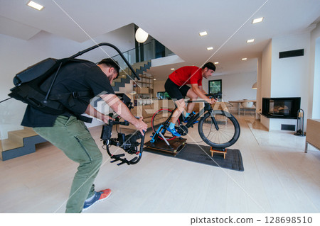 A cameraman filming an athlete riding a triathlon bike on a simulation machine in a modern living room. Training in pandemic conditions A cameraman filming an athlete riding a triathlon bike on a simulation machine in a modern living room. Training in pandemic conditions 128698510