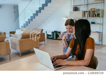 Mother with her daughter talking on laptop with family and friends while sitting in modern living room of big house. Mother with her daughter talking on laptop with family and friends while sitting in modern living room of big house. 128698539