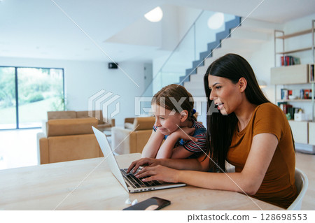 Mother with her daughter talking on laptop with family and friends while sitting in modern living room of big house. Mother with her daughter talking on laptop with family and friends while sitting in modern living room of big house. 128698553