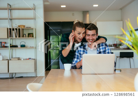 A young married couple is talking to parents, family and friends on a video call via a laptop while sitting in the living room of their modern house. 128698561