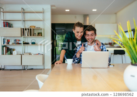 A young married couple is talking to parents, family and friends on a video call via a laptop while sitting in the living room of their modern house. 128698562
