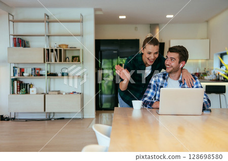 A young married couple is talking to parents, family and friends on a video call via a laptop while sitting in the living room of their modern house. 128698580