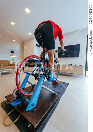 A triathlete riding a triathlon bike on a machine simulation in a modern living room. Training during pandemic conditions. 128698593