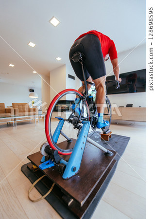 A triathlete riding a triathlon bike on a machine simulation in a modern living room. Training during pandemic conditions. 128698596