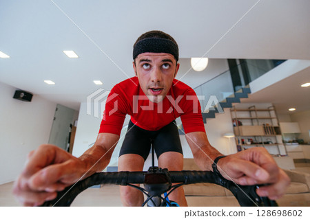 A triathlete riding a triathlon bike on a machine simulation in a modern living room. Training during pandemic conditions. A triathlete riding a triathlon bike on a machine simulation in a modern living room. Training during pandemic conditions. 128698602