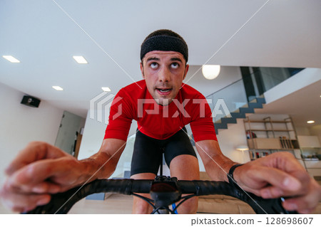 A triathlete riding a triathlon bike on a machine simulation in a modern living room. Training during pandemic conditions. 128698607