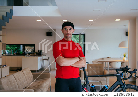 Photo of a determined triathlete standing in a modern large living room, behind him is his training bike 128698612