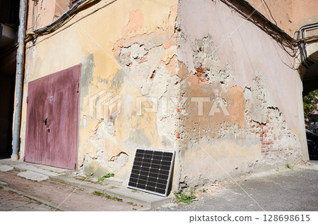 Photovoltaic solar panel at weathered, textured wall with peeling paint. Juxtaposition of modern renewable energy technology against rustic, decaying backdrop highlights contrast between old and new. 128698615