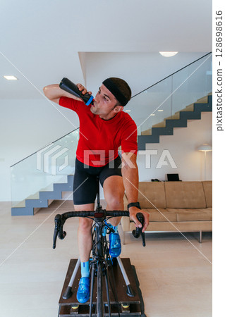 An athlete drinking water while riding a triathlon bike on a simulation machine in a modern living room. Training in pandemic conditions 128698616