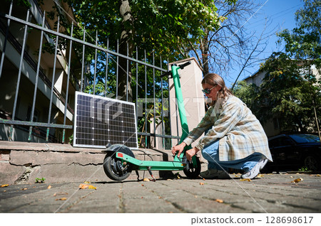 Woman connecting solar panel to electric scooter, highlighting practical application of sustainable renewable energy. Concept of integration of eco-friendly technology in everyday transportation. Woman connecting solar panel to electric scooter, highlighting practical application of sustainable renewable energy. Concept of integration of eco-friendly technology in everyday transportation. 128698617