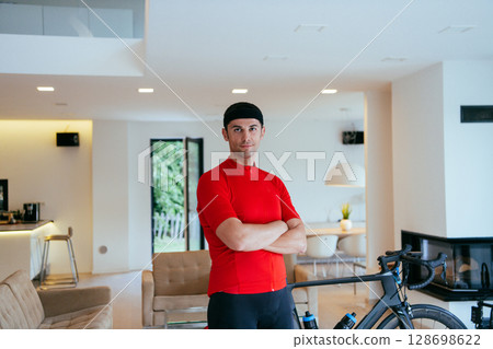 Photo of a determined triathlete standing in a modern large living room, behind him is his training bike 128698622