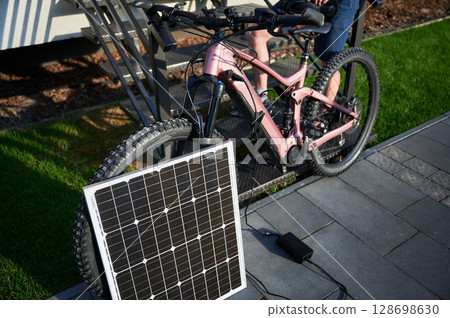 Pink electric mountain bike parked beside staircase, connected to small solar panel for charging. Bike rugged tires, situated on paved pathway with grassy lawn. Pink electric mountain bike parked beside staircase, connected to small solar panel for charging. Bike rugged tires, situated on paved pathway with grassy lawn. 128698630