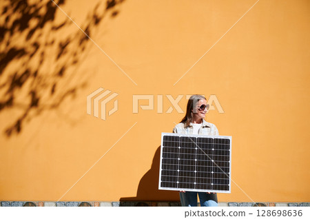 Woman holding solar panel for charging, stands against vibrant orange wall. Smiling girl wears sunglasses, white top. Artistic shadows cast by nearby trees branches on wall. 128698636