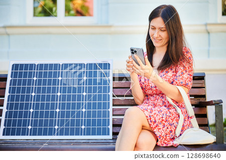 Pretty woman charging her phone from solar panel. Young female using portable solar battery to charge her device. Alternative sources of energy, saving environment concept 128698640