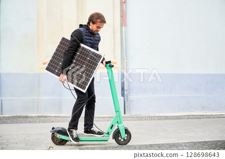 Man rides electric scooter while carrying solar panel. Integration of solar power as sustainable energy source for charging electric scooters, promoting eco-friendly urban transportation. 128698643