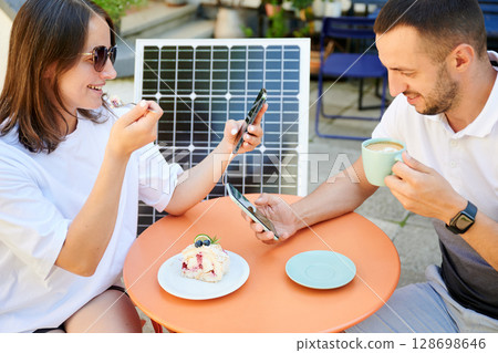 Man and woman charging smartphone from solar panel, relaxing in cafe. Cute couple using portable solar charger for devices. Charming couple sitting on terrace on background of solar battery. 128698646