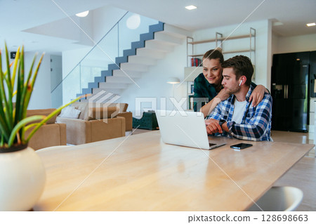 A young married couple is talking to parents, family and friends on a video call via a laptop while sitting in the living room of their modern house. A young married couple is talking to parents, family and friends on a video call via a laptop while sitting in the living room of their modern house. 128698663