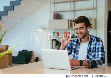 Businessman sitting at a table in a modern living room, with headphones using a laptop for business video chat, conversation with friends and entertainment. 128698672