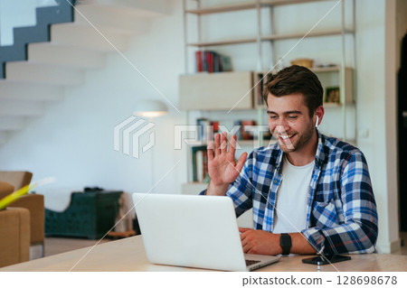 Businessman sitting at a table in a modern living room, with headphones using a laptop for business video chat, conversation with friends and entertainment. 128698678