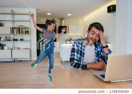 Work from home. Daughter interrupting her father while he is having a business online conversation on his laptop while sitting in the modern living room. 128698690