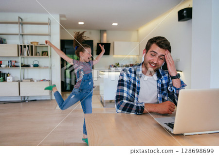 Work from home. Daughter interrupting her father while he is having a business online conversation on his laptop while sitting in the modern living room. Work from home. Daughter interrupting her father while he is having a business online conversation on his laptop while sitting in the modern living room. 128698699