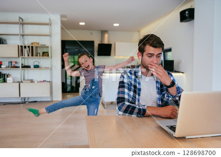 Work from home. Daughter interrupting her father while he is having a business online conversation on his laptop while sitting in the modern living room. 128698700