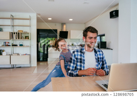 Work from home. Daughter interrupting her father while he is having a business online conversation on his laptop while sitting in the modern living room. 128698705
