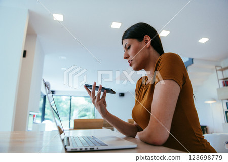 A woman sitting in the living room using a laptop and a smartphone look at camtalk by video call with business friends and relatives, head shot. Job interview answering questions.  128698779