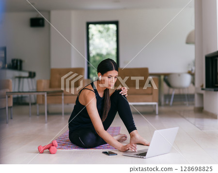 A woman in sportswear is sitting in the living room and preparing for online training while using a laptop. 128698825