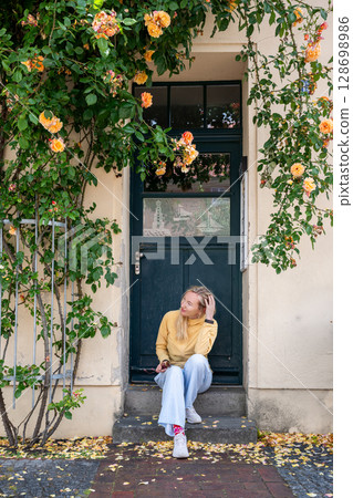 Young woman sitting on doorstep under blooming rose bush in front of vintage house facade 128698986