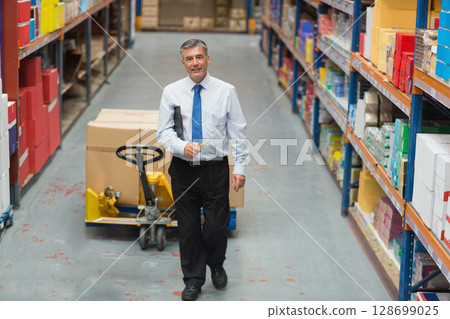 Walking senior man holding clipboard and pencil in warehouse aisle, with yellow pallet jack 128699025