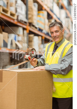 Man in his 50s sealing cardboard box with tape dispenser in warehouse aisle, copy space Man in his 50s sealing cardboard box with tape dispenser in warehouse aisle, copy space 128699027