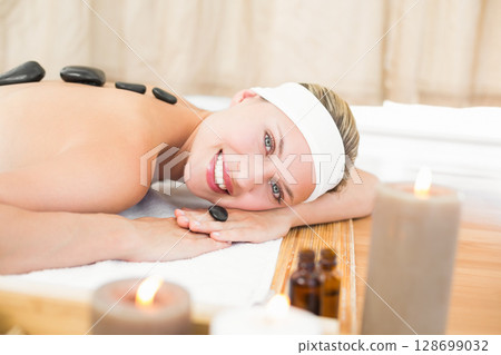 Relaxing female client lying on massage table in spa treatment room, with hot stones and candles 128699032